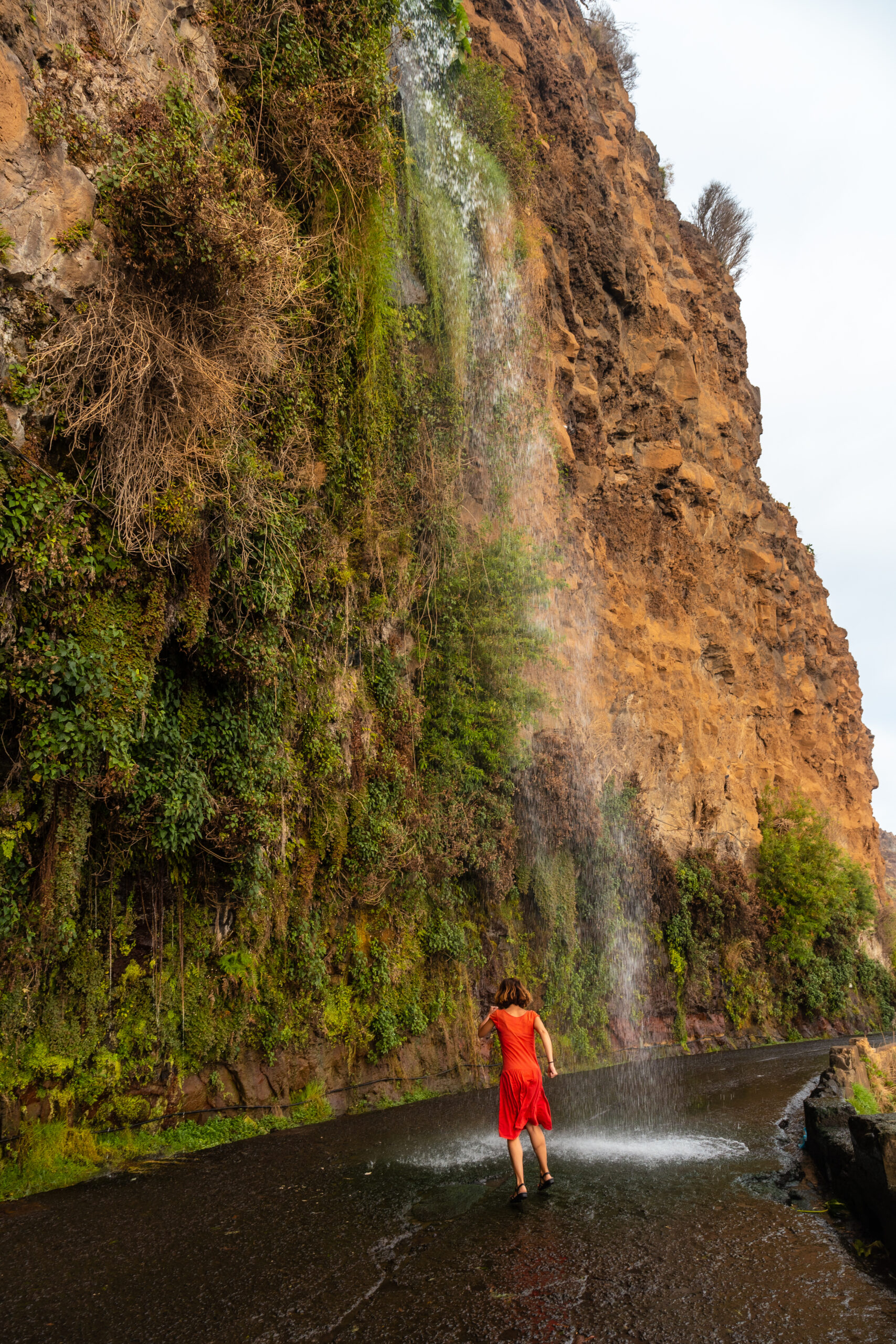 A young woman in a red dress at the waterfall that falls on the road called Anjos Waterfall, Madeira